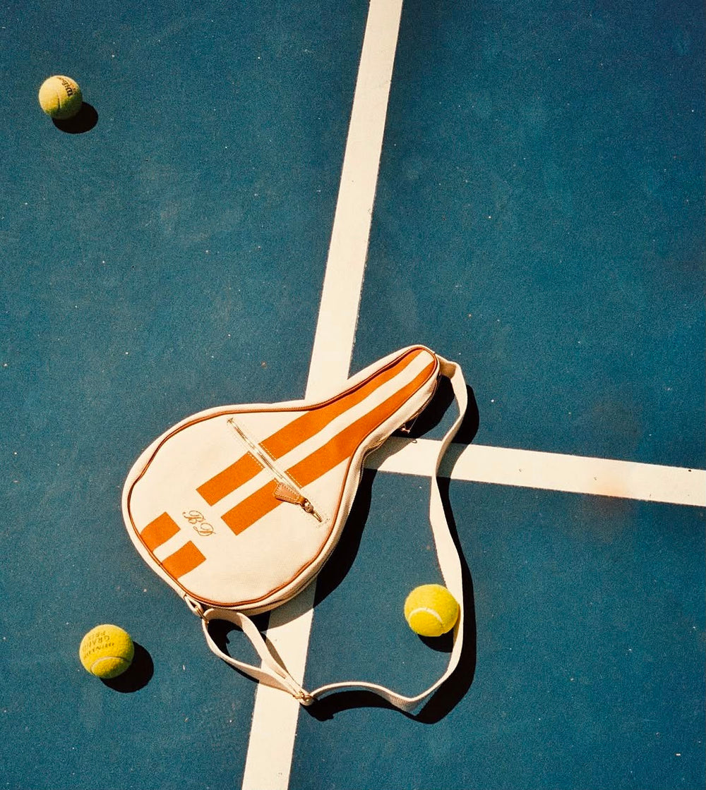 A tennis bag with orange and white stripes lies on a blue tennis court, crossed by a white boundary line, with three yellow tennis balls around it.